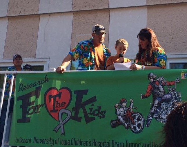 Matthew Wessels speaking into a microphone at the ride kickoff, beside the Research For The Kids banner