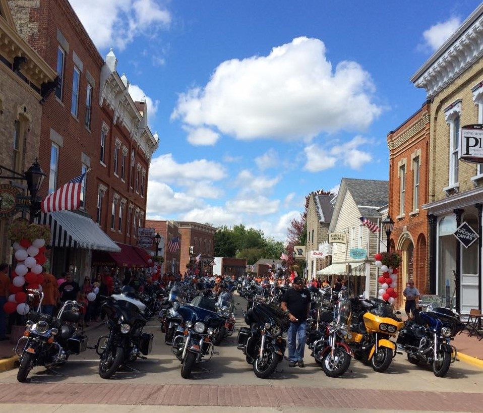 A small-town main street lined on both sides with parked motorcycles under a blue sky, storefronts and flags behind them